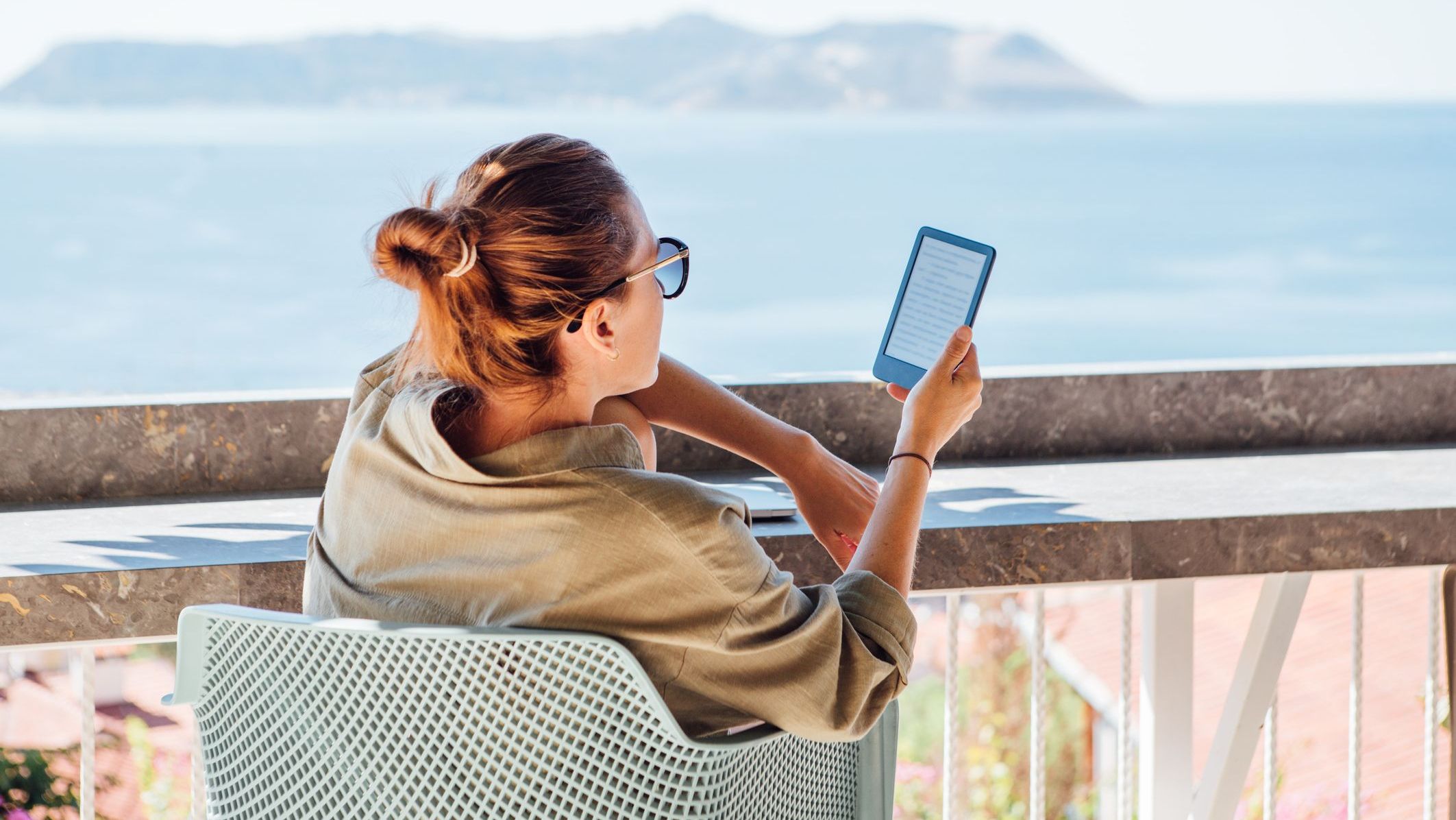 Middle-aged beautiful woman relaxing with e-book on sunny house terrace lit by morning sunlight, overlooking Mediterranean Sea harbor