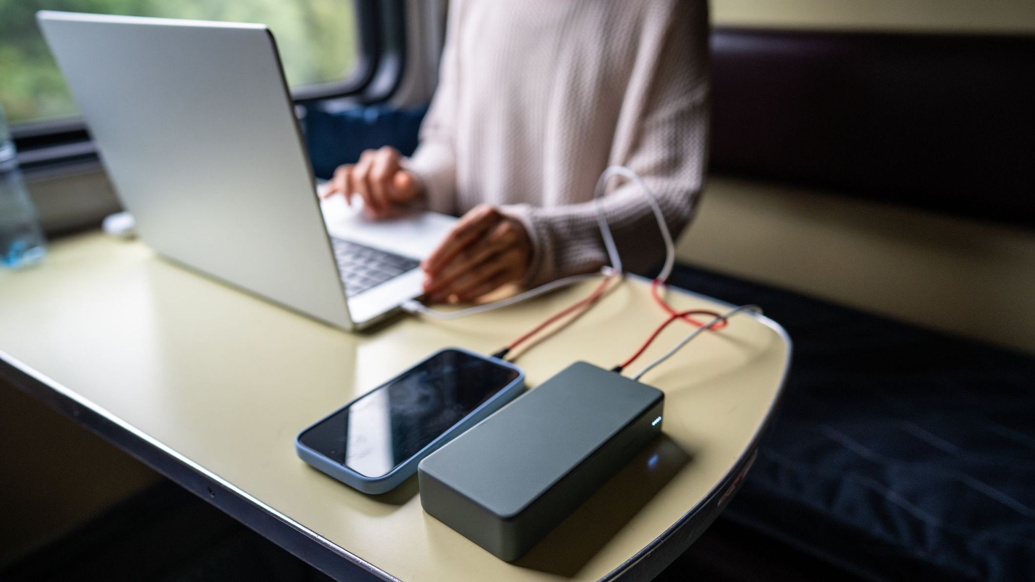 Woman working on laptop, having it and phone connected to power bank, sitting in train compartment.