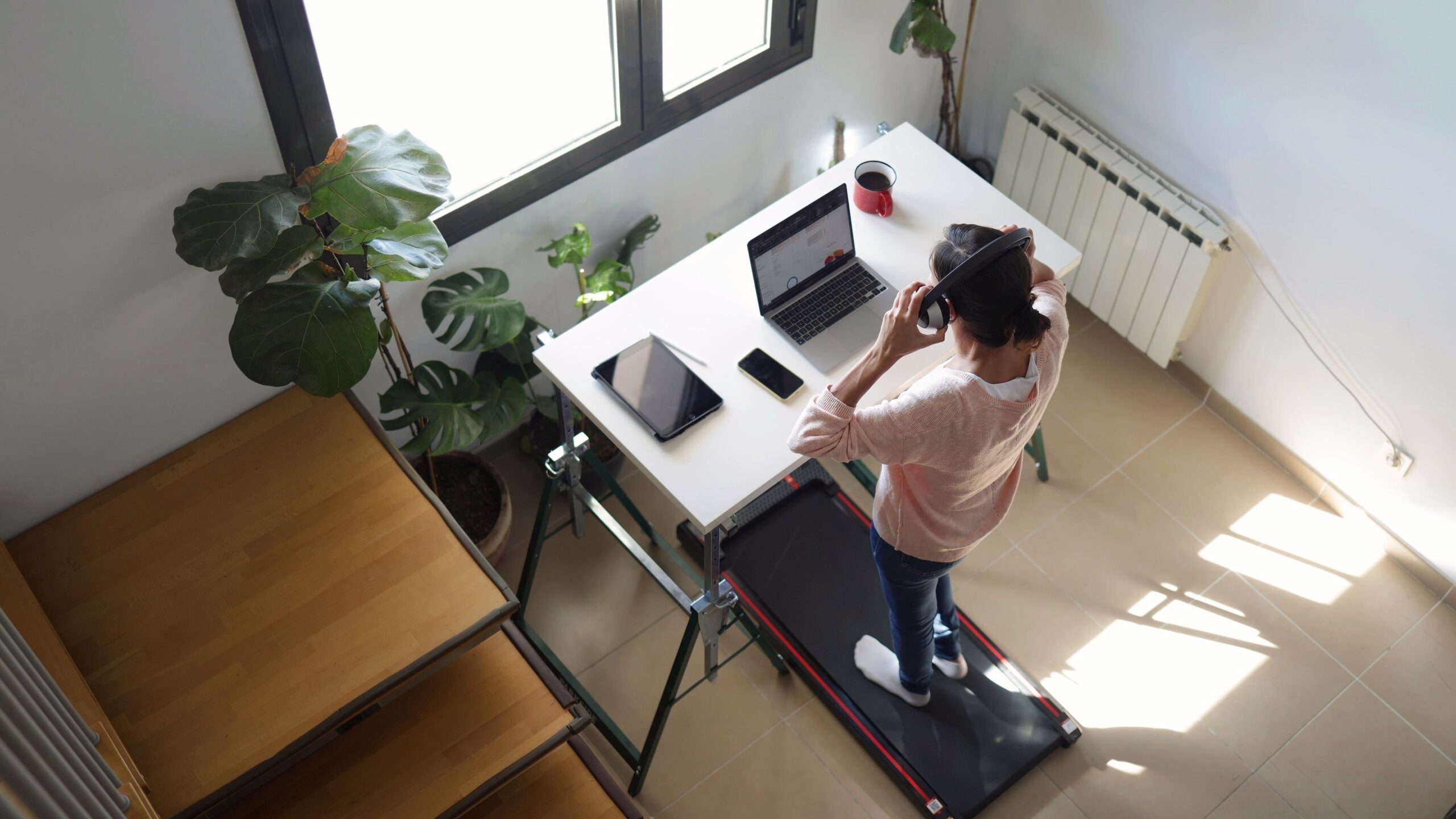Woman working from home at a standing desk with a laptop, putting on headphones, doing light exercise on a walking pad, prioritizing health