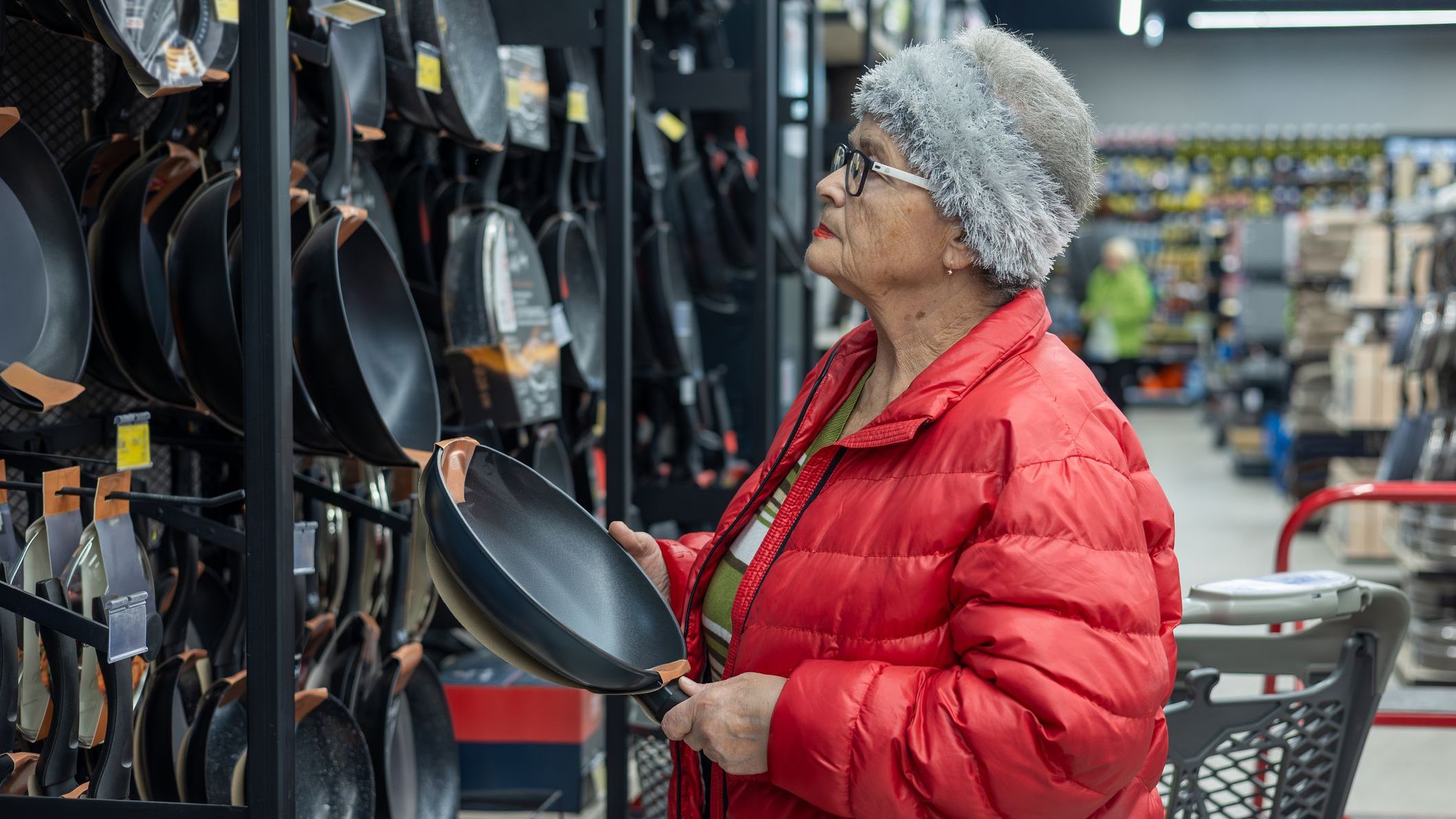An elderly woman shopping independently in a supermarket housewares section, carefully choosing a frying pan from a wide selection of cookwa