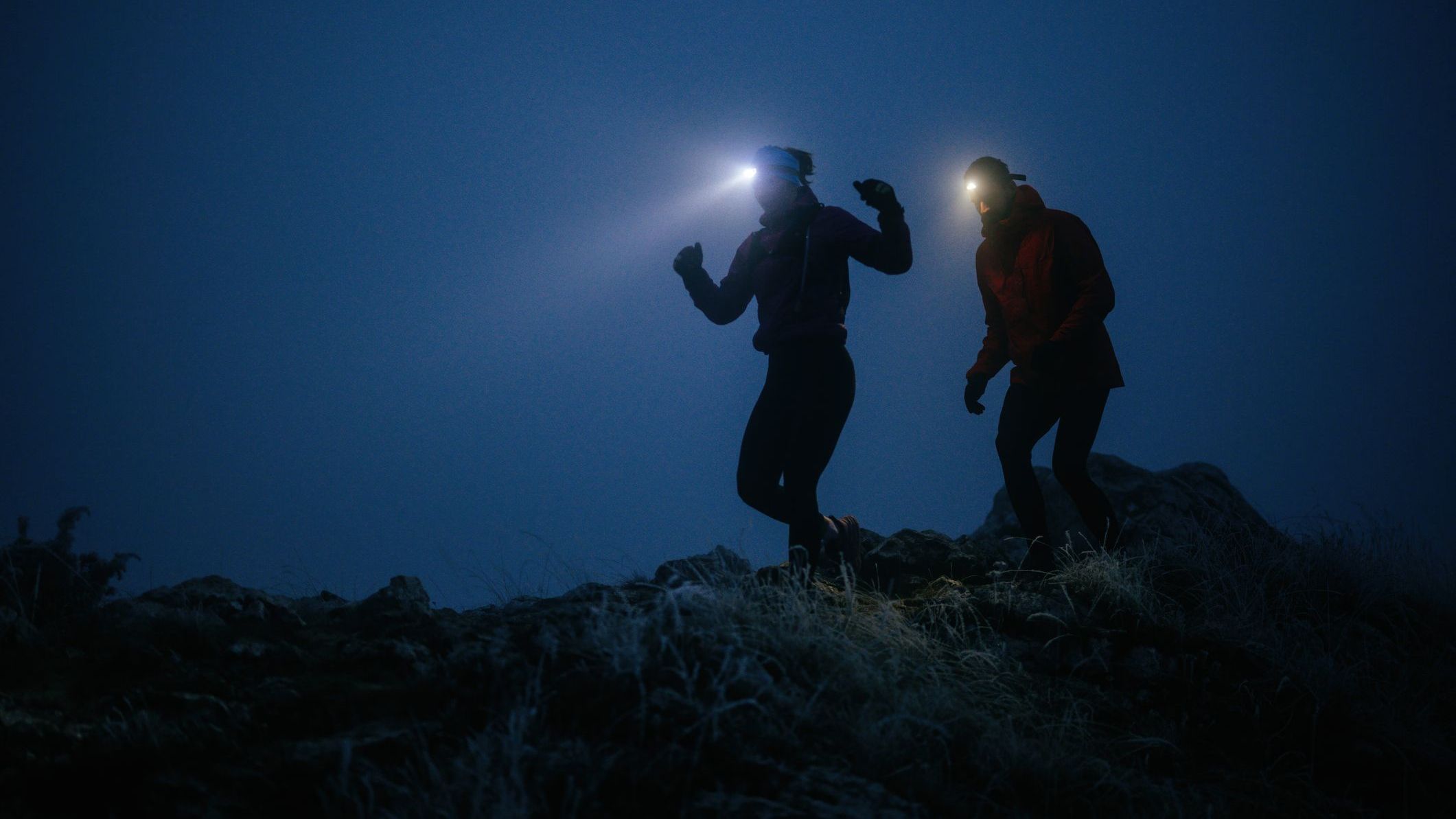 Two athletes run along a rugged mountain trail at night, wearing headlamps and winter gear, navigating the cold, misty terrain with determin