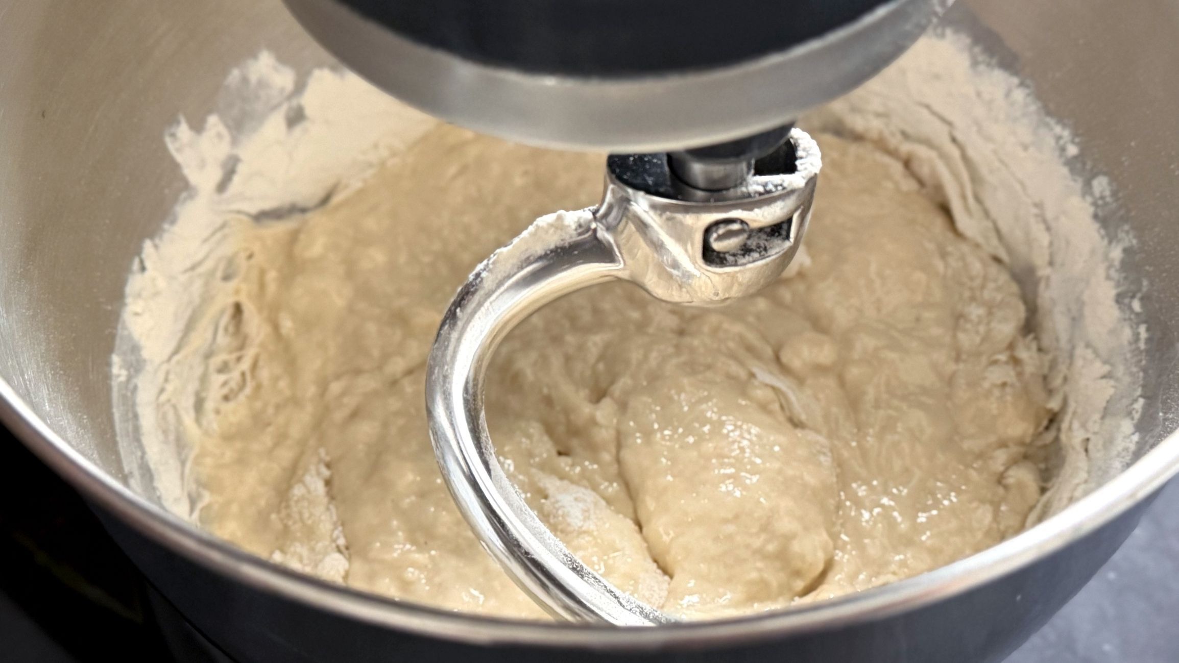 Stand mixer with a dough hook blends flour and water into a smooth bread dough inside a stainless steel bowl on a kitchen countertop