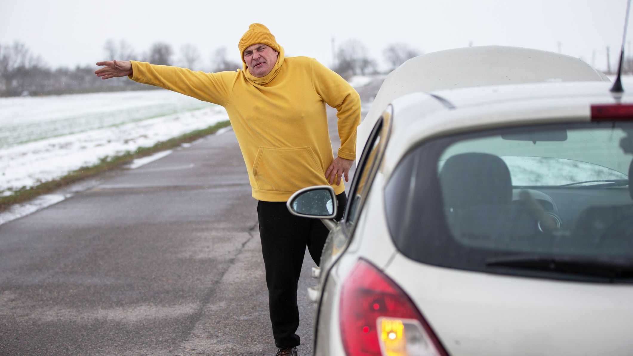 A man wearing a yellow hoodie stands next to his car with the hood raised, signaling for assistance on a snowy rural road. The overcast sky