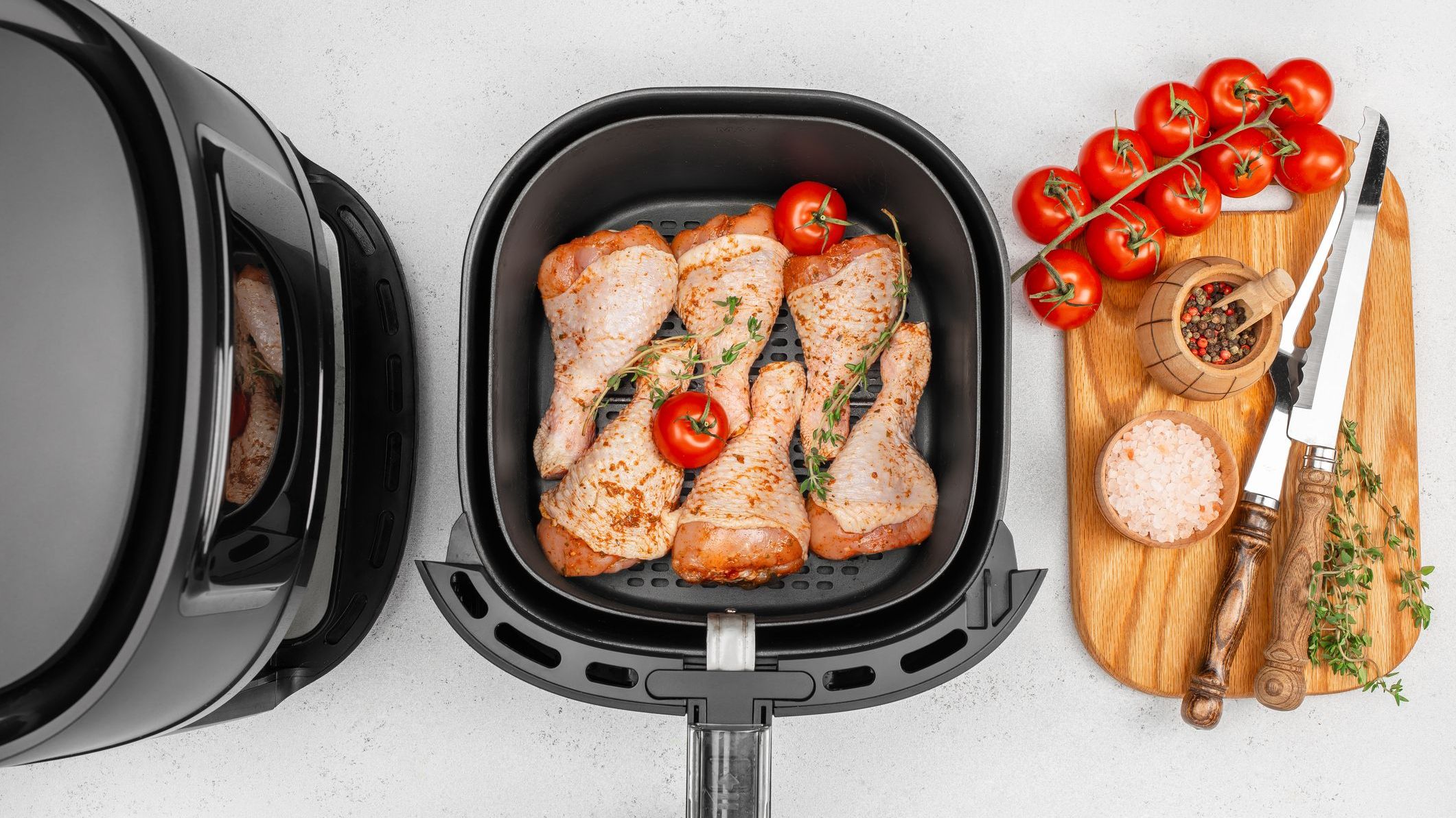 Raw seasoned chicken drumsticks with fresh tomatoes and herbs are arranged in an air fryer basket, surrounded by spices, cherry tomatoes on