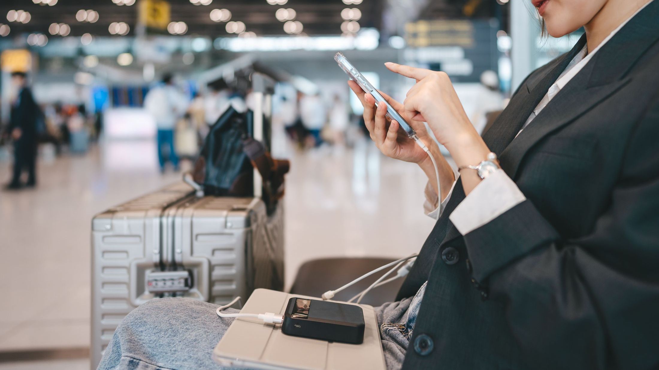 Young asian business woman use smartphone device charging mobile with power bank at international airport terminal departure platform
