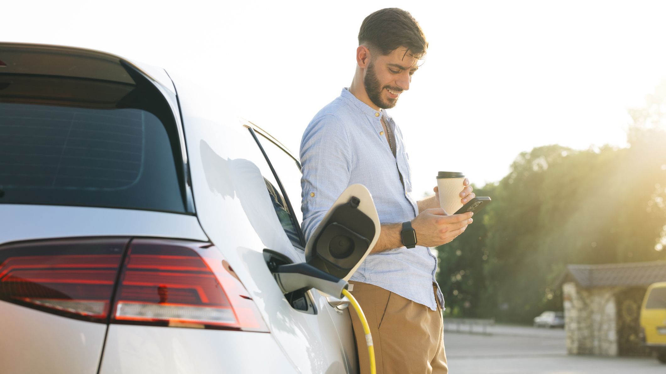 Gorgeous bearded caucasian man in a blue shirt standing near an electric car that is charging